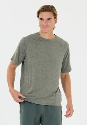 Young man with short light brown hair wearing a textured grey short-sleeve shirt and dark grey pants, standing against a plain background.