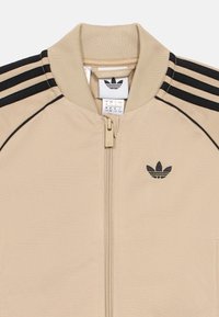 Bež jakna na patentni zatvarač s rebrastom ovratnikom, tri crne pruge na ramenima i malim crnim logotipom na prsima. Glatka tekstura tkanine.