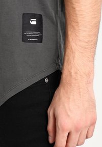 Šedé tričko s oblým lemem, které má černý logo nášivku na levé straně a minimální švy. Nosit s černými džíny.