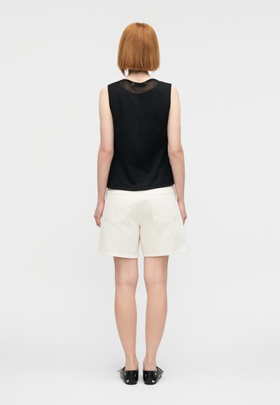 Haut noir sans manches avec un col en tulle transparent, associé à un short blanc avec deux poches arrière. Le modèle porte des chaussures plates noires.