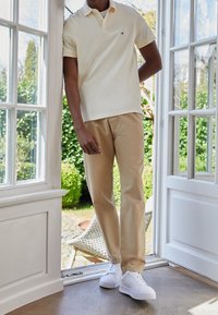 Vit pikétröja, beige chinos och vita sneakers. Enkelt mönster, kort ärm och klassisk passform, står bredvid en dörröppning med grönska.