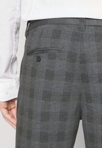 Grå rutig byxor i texturerad tyg med ett subtilt rutmönster. Har en bakficka med en svart knappdetalj.