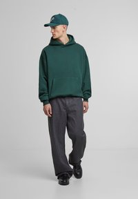 Felpa verde con tasca a marsupio, abbinata a jeans neri larghi e scarpe nere. Il cappellino è verde abbinato con logo ricamato.