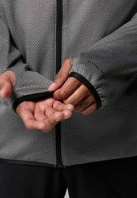 Szara kurtka zapinana na zamek z teksturowanej powierzchni, z dopasowanymi mankietami z otworami na kciuki i czarnymi wykończeniami, przedstawiająca ręce poprawiające rękawy.