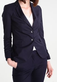 Blazer bleu marine en tissu texturé, avec un col cranté, deux boutons et deux poches avant. Associé à un pantalon assorti.