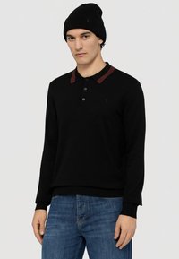 Černé polotričko s dlouhým rukávem a bordovým límečkem, vybavené tříknoflíkovou légou a žebrovanými manžetami, ve spojení s modrými džínami.