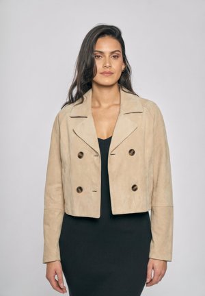 Donna con lunghi capelli scuri che indossa una giacca beige doppiopetto corta sopra un vestito nero a coste, in piedi davanti a uno sfondo chiaro e uniforme.