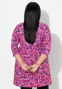 Kwiecista tunika w różu, fiolecie i czerni z rękawami do połowy ramienia, wymarszczoną talią i luźnym krojem, noszona na czarnych legginsach.