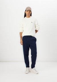 Hvit kabelstrikket genser, marineblå joggebukser med blå sidestrekk, hvite sneakers og en beige caps. Enkelt design med ribbede teksturer.
