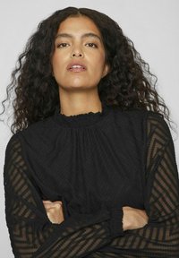 Black blouse with textured, sheer long sleeves. Features a ruffled mock neck and zigzag pattern, creating a layered appearance.
