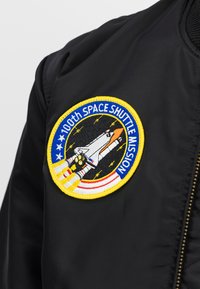 Giacca nera con una toppa rotonda ricamata che mostra il lancio di uno space shuttle e il testo "100a MISSIONE DELLO SPACE SHUTTLE" in colori blu e giallo.