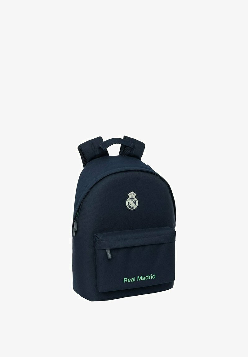 Sac à dos bleu marine avec des bretelles rembourrées, poche avant, et texte vert "Real Madrid". Il présente un logo circulaire en haut.