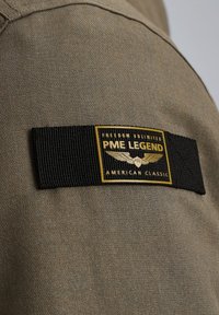 Beżowa tkanina z teksturowaną powierzchnią zawiera czarną, wplecioną metkę z złotym napisem i symbolem skrzydła, mówiącym "PME LEGEND AMERICAN CLASSIC."