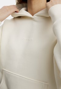 Krämfärgad hoodie med en mjuk textur, med en framficka och en liten präglad logotyp. Huvtröja med ribbade detaljer.