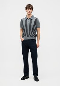 Νέος άνδρας που στέκεται με το χέρι στην τσέπη, φορώντας γκρι και μαύρο ριγέ κοντομάνικο πόλο μπλουζάκι, μαύρο παντελόνι και μαύρα παπούτσια σε λευκό φόντο.