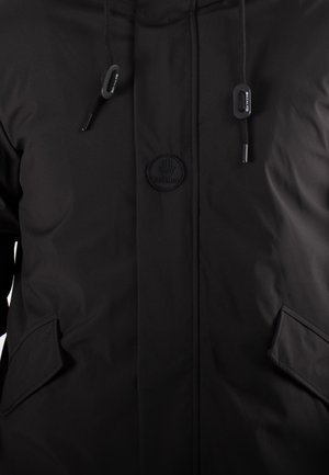 Veste matelassée noire avec une texture douce, un logo rond, des cordons de serrage réglables et deux poches latérales à rabat.