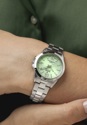 Orologio da polso con quadrante verde, cinturino in metallo argento, che segna le 3:40 e la data 20, indossato al polso di una persona su uno sfondo di camicia verde.