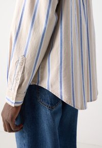 Beige langwerpige shirt met blauwe verticale strepen. Heeft een knoopmanchet, een ontspannen pasvorm en een licht gebogen zoom. Gepaard met blauwe denim jeans.