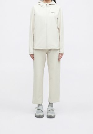 Osoba oblečená v krémové bundě na zip s kapucí a odpovídajících kalhotách, šedých ponožkách a šedých teniskách se suchým zipem, stojící na bílém pozadí.