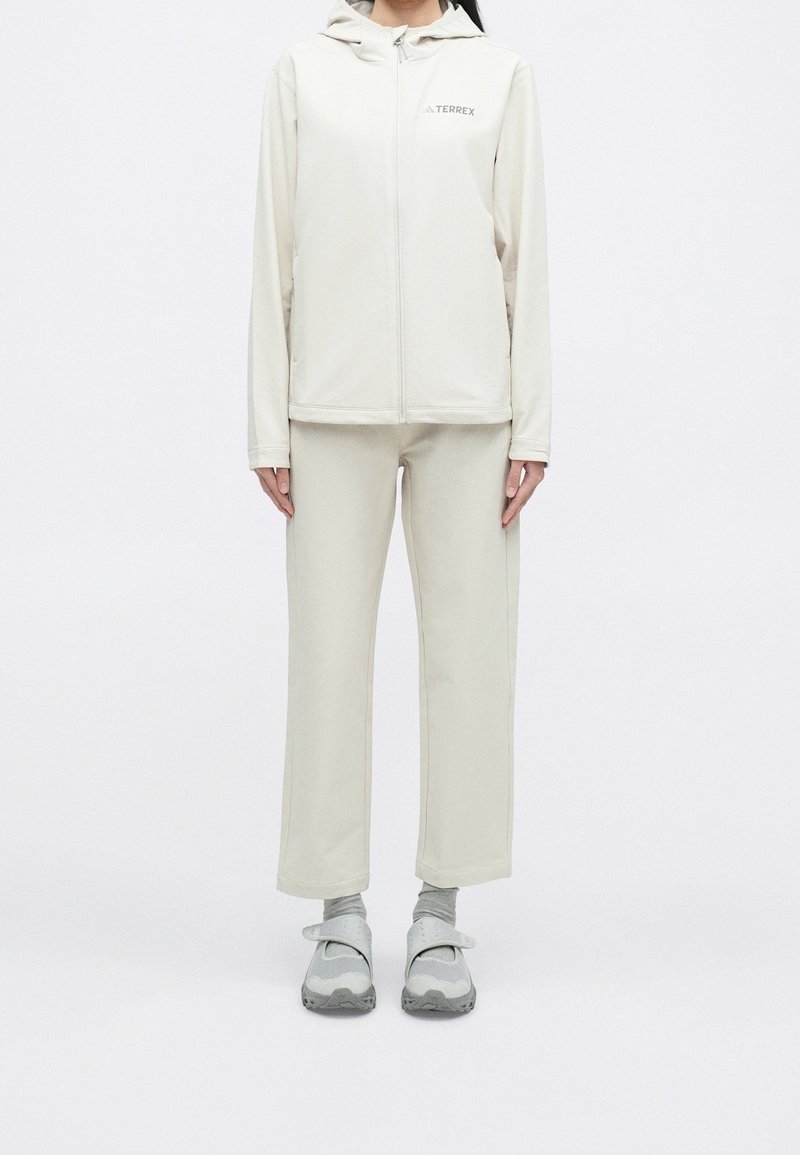 Person trägt eine cremefarbene, mit Reißverschluss versehene Kapuzenjacke und passende Hosen, graue Socken und graue Sneakers mit Riemen, steht vor weißem Hintergrund.