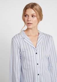 Pyjamatopp i ljusblått med vertikala marinblå ränder. Har en skärning vid kragen, långa ärmar och knappstängning framtill med marinblå knappar.