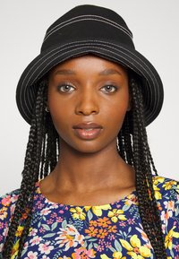 Chapéu estilo bucket preto com costura branca, feito de algodão, com uma aba larga. O modelo usa uma blusa com padrão floral em cores vibrantes.