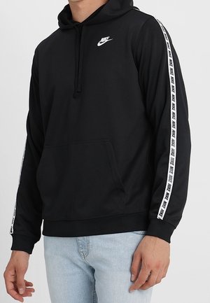 Mand iført sort Nike hoodie med hvidt logo på brystet og brandede striber ned ad ærmerne, kombineret med lyseblå jeans.