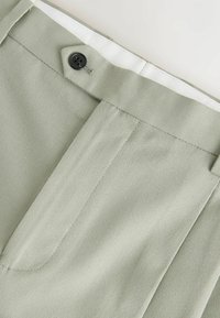 Gros plan sur la ceinture d’un pantalon gris clair avec un bouton noir et une boucle de ceinture visible, montrant le tissu texturé et les détails des coutures.