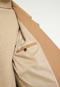 Beige uldfrakke med en struktureret overflade, der har en glat inderforing og en lille, knaplukket lomme på venstre side.