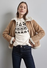 Tan-suede-takki, jossa on valkoinen keinoturkiskaulus ja -hiharesorit, päällä kerma Beige neule, jossa on mustat geometriset kuviot, yhdistettynä tummiin farkkuihin.