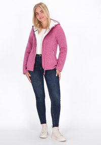 Chaqueta de forro polar rosa con interior blanco, cierre de cremallera en la parte delantera y capucha ajustable, combinada con jeans oscuros y zapatillas de color claro.