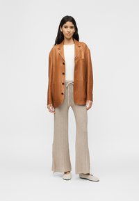 Γυναίκα στέκεται φορώντας καφέ δερμάτινο σακάκι πάνω από λευκό ριμπ πλεκτό τοπ, μπεζ παντελόνι πλεκτό με φαρδιά μπατζάκια και κρεμ παντόφλες σε απλό φόντο.