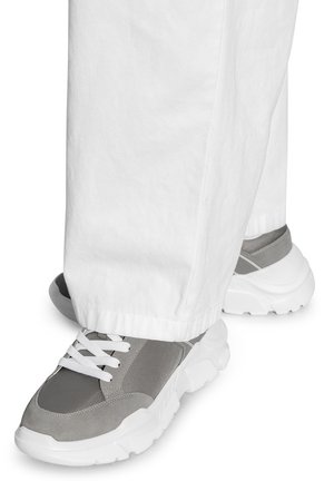 Des baskets athlétiques grises avec des lacets blancs, présentant un mélange de matériaux en daim et en maille, et une semelle en caoutchouc blanc épais. Portées avec un pantalon blanc.
