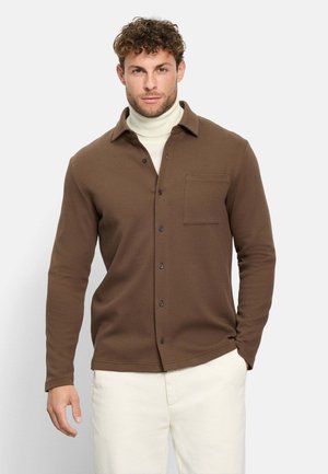 Mann trägt braunes Hemd mit Knöpfen über einem weißen Rollkragenpullover und hellen Hosen, steht vor schlichtem hellem Hintergrund.