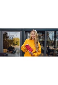 Red wallet with a button closure, made of leather, held in hands. The background features large windows and soft furnishings in neutral tones.