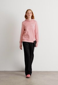 Sudadera rosa clara con un bolsillo frontal, pantalones anchos negros y zapatos rosas. Tejido suave y corte relajado con un detalle de logo en el pecho.