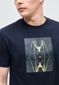 Άνδρας που φοράει μπλε ναυτικό t-shirt με κίτρινο αφηρημένο σχέδιο αράχνης και το κείμενο "BOSS" στο κέντρο του στήθους.