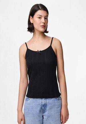 Jeune femme aux cheveux courts et foncés, portant un top noir sans manches à fines bretelles et un jean bleu clair, debout sur fond blanc.