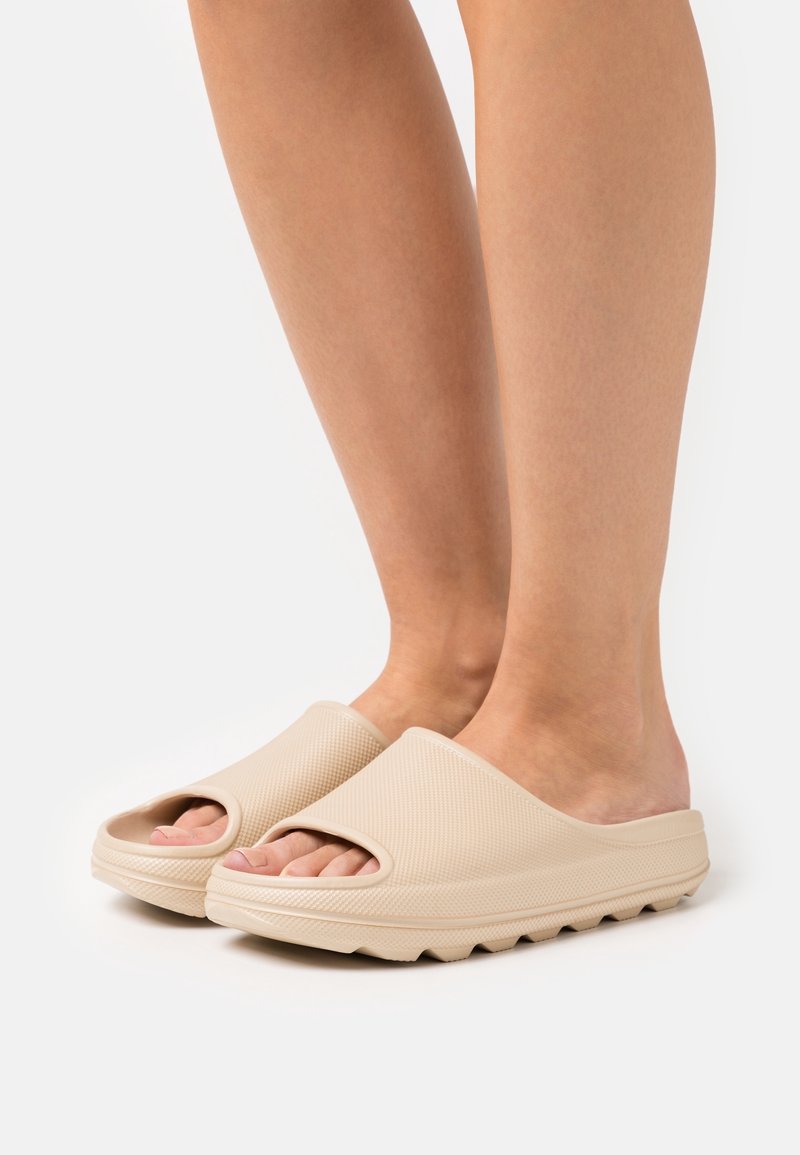 Pieds portant des chaussons beige texturés, à enfiler, à bout ouvert, avec des semelles épaisses et des dessous rainurés sur fond blanc.