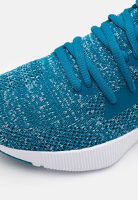 Zapatilla deportiva en color verde azulado con parte superior de punto texturizado, acentos en gris claro y suela de goma blanca. Cuenta con cordones planos y un collar acolchado.