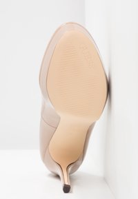 Chaussure beige à talon haut avec bout pointu, montrant la semelle avec une zone de grip texturée et le logo "GUESS", inclinée contre un coin blanc.