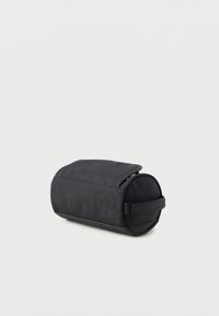 Black cylindrical bag made of smooth fabric with a flap closure, featuring a textured side and a logo-embossed handle.