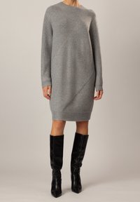 Robe en tricot grise avec un design diagonal, des manches longues et des accents côtelés. Portée avec des bottes en cuir noir jusqu'aux genoux.