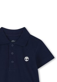 Polo de color azul marino hecho de tejido texturizado, con un cuello de tres botones y un logo de árbol blanco en el lado izquierdo del pecho.
