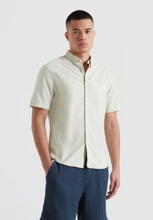 Mann trägt ein hellbeiges kurzärmeliges Hemd mit Knöpfen und strukturierte marineblaue Hose, steht mit einer Hand in der Tasche vor weißem Hintergrund.