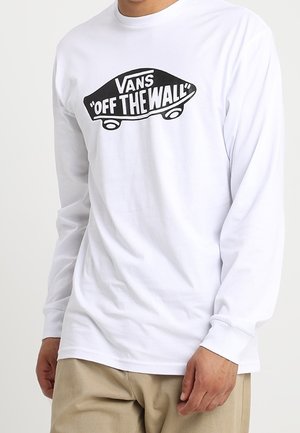 Person trägt ein weißes Langarmshirt mit schwarzem "VANS OFF THE WALL" Skateboard-Logo auf der Vorderseite, kombiniert mit beigen Hosen.