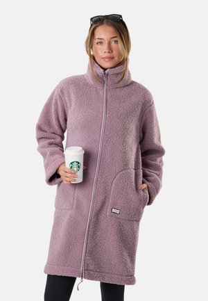 Kobieta o jasnobrązowych włosach, z okularami przeciwsłonecznymi na głowie, ubrana w długi fioletowy polar, trzymająca w prawej ręce kubek Starbucks.