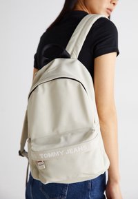 Ung person som bär en krämfärgad Tommy Jeans ryggsäck med vadderade remmar över en svart tröja och blå jeans, vänd bort från kameran.