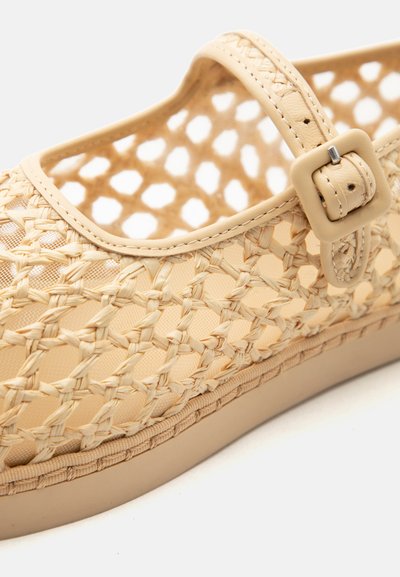 Šviesiai rudos spalvos pynė plokščias batai su reguliuojamu dirželiu ir sagtimi, su tekstūruota padu ir atviru tinklinio dizainu viršuje.