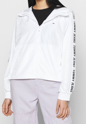 Persona con una chaqueta blanca con cremallera y texto "Tommy Jeans" en las mangas y pantalones de color lila claro sobre un fondo liso.